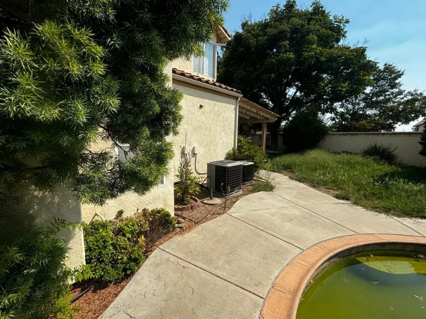A two-story stucco house with a path, overgrown yard, and a round, green-tinted pool.