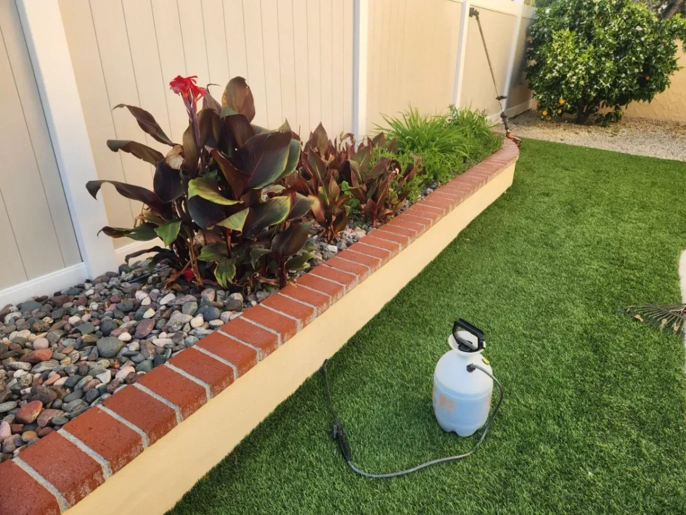 A flower bed with red brick and beige border next to artificial turf and a blue sprayer.