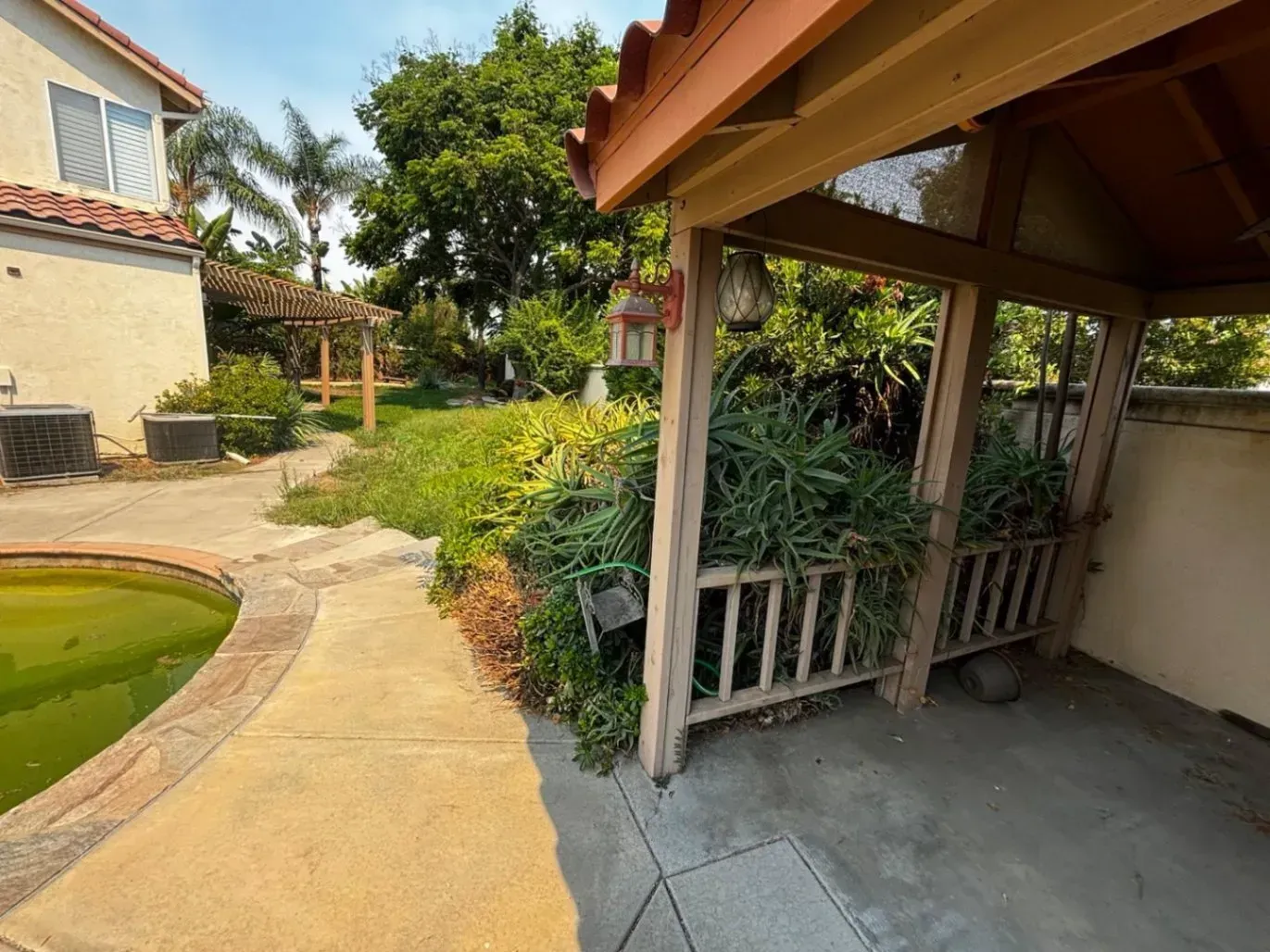 Backyard scene with a pool, patio, and covered seating area. Lush greenery and a house.
