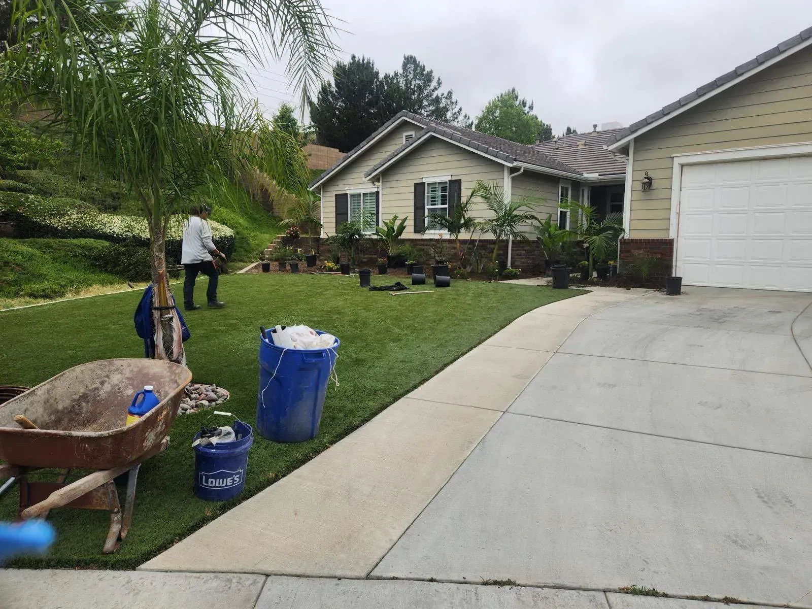 Lawn care: person planting trees in a yard with a house and driveway. Equipment and supplies are present.