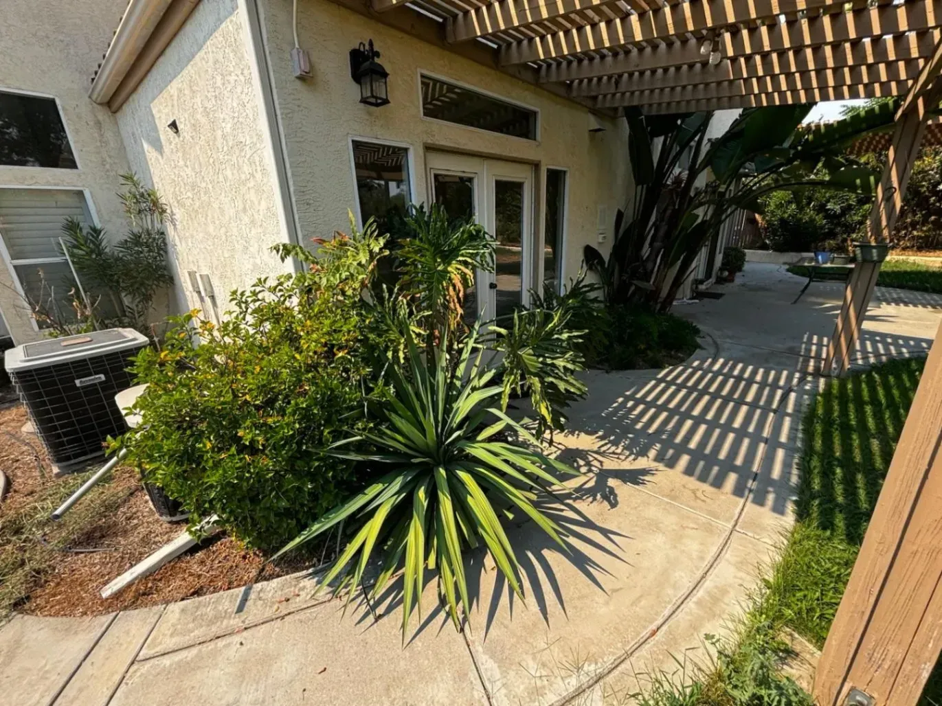 A building exterior with a curved walkway, a pergola, and greenery. An air conditioning unit is visible.