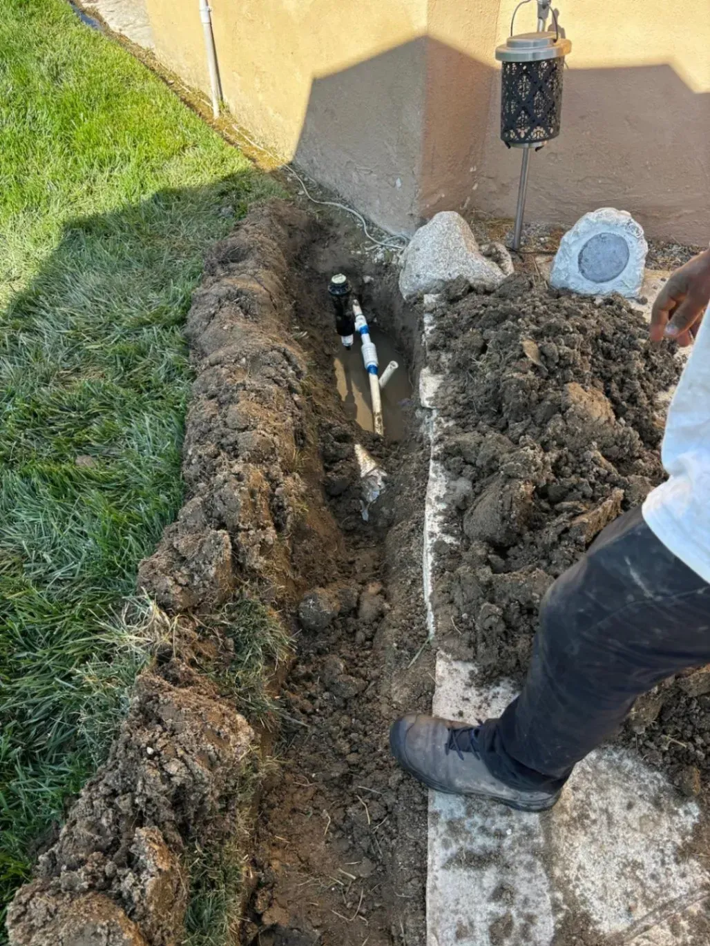 A trench dug in a yard reveals exposed pipes. A person stands nearby.