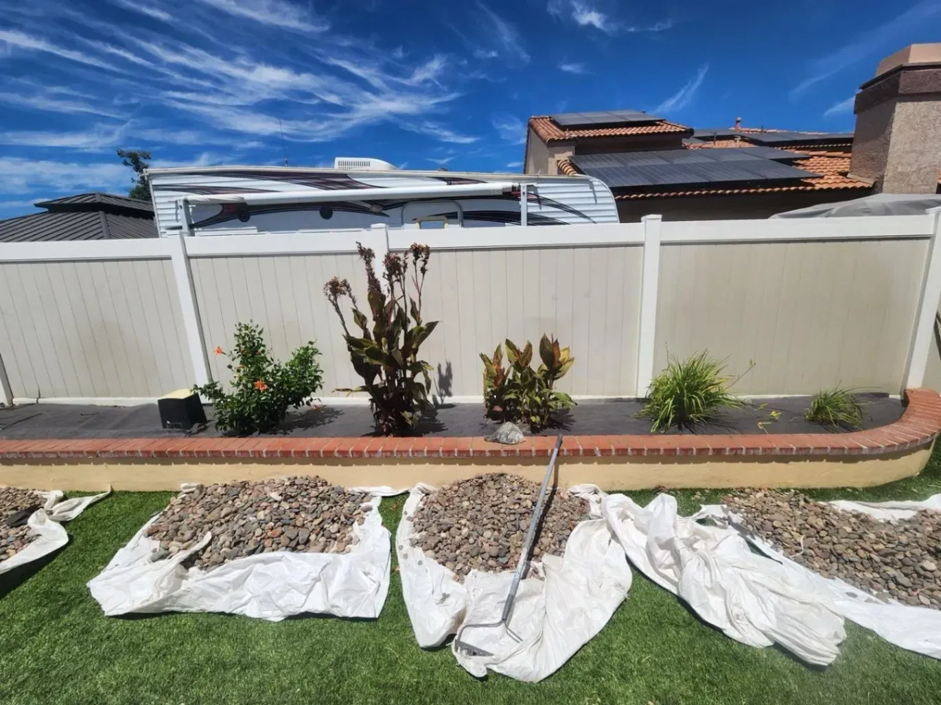 Yard work scene: mulch in bags on grass, plants in front of a fence, RV visible, sunny day.