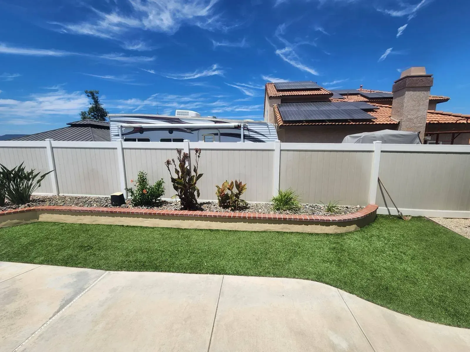 Backyard with beige fence, green lawn, flower bed, and blue sky. House with solar panels visible.
