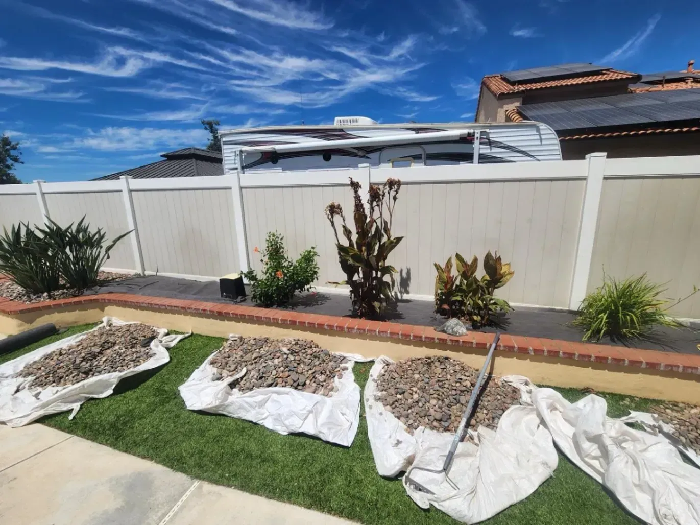 A backyard with a white fence, landscaping, and bags of gravel on artificial turf under a blue sky.