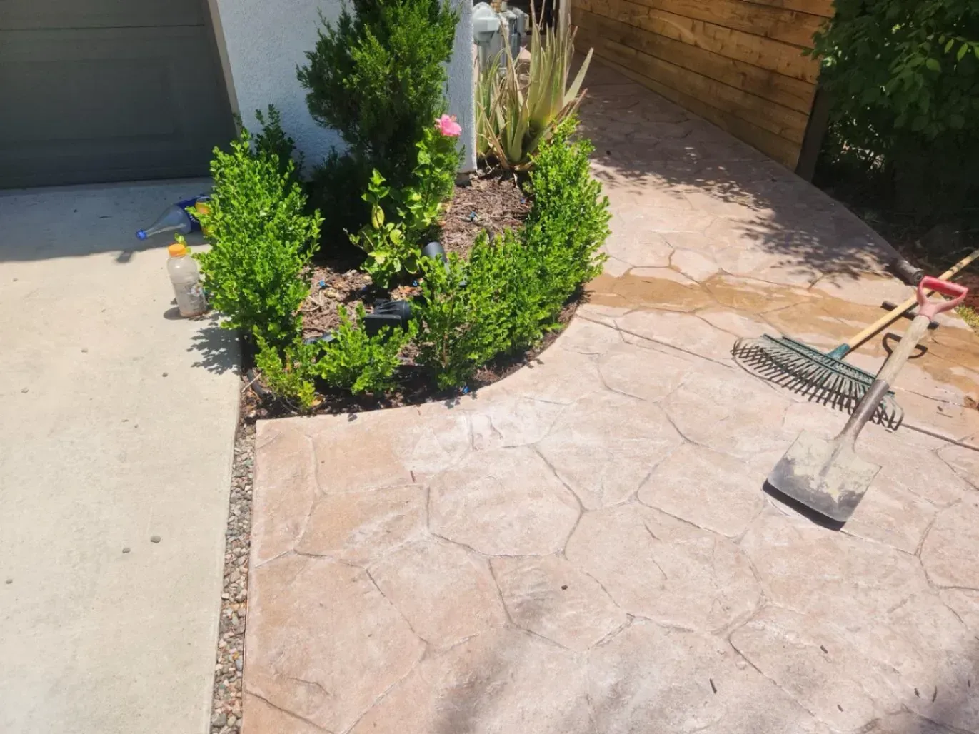 Landscaped area with brown hexagonal stamped concrete and green bushes surrounding a flower bed. A rake leans against the area.