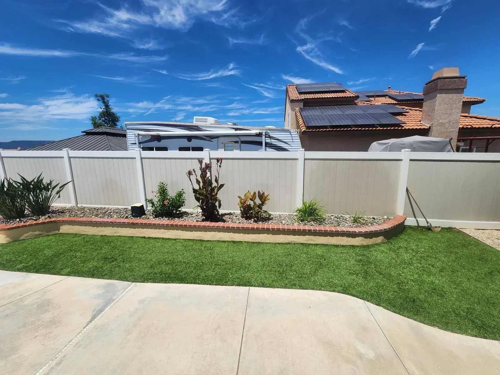 Lawn with a small brick-bordered garden in front of a neutral-colored fence with a house behind it under a blue sky.