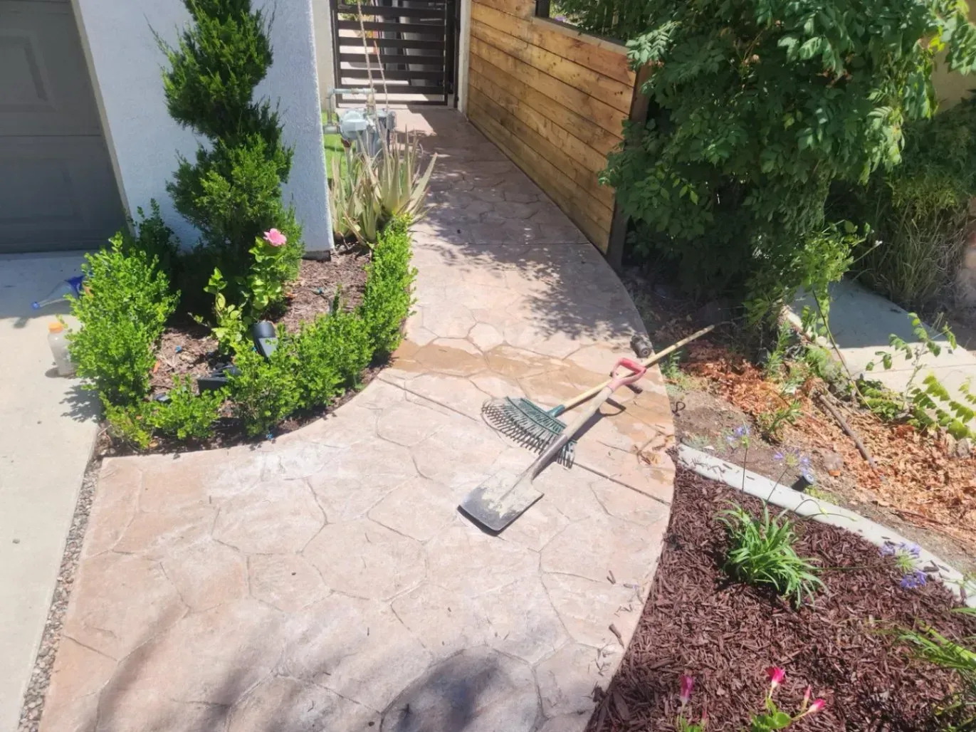 Pathway with stamped concrete, surrounded by landscaping, with shovel and rake lying on the path.