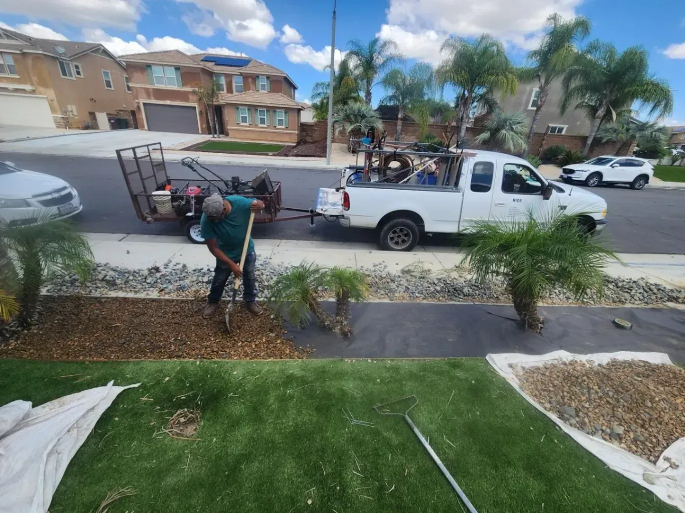 Man shoveling mulch near a lawn with a truck and trailer on a street in front of houses.