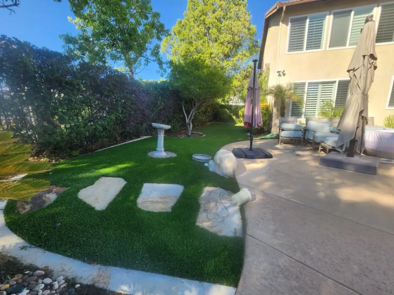 Backyard with green lawn, stepping stones, birdbath, trees, and house with patio furniture.