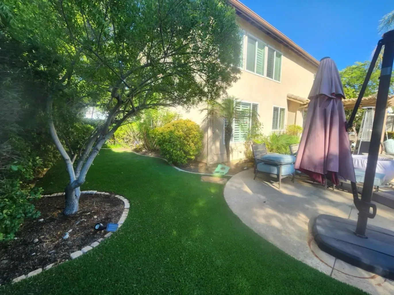 Backyard with green artificial turf, a tree, patio furniture, and a house under a sunny sky.
