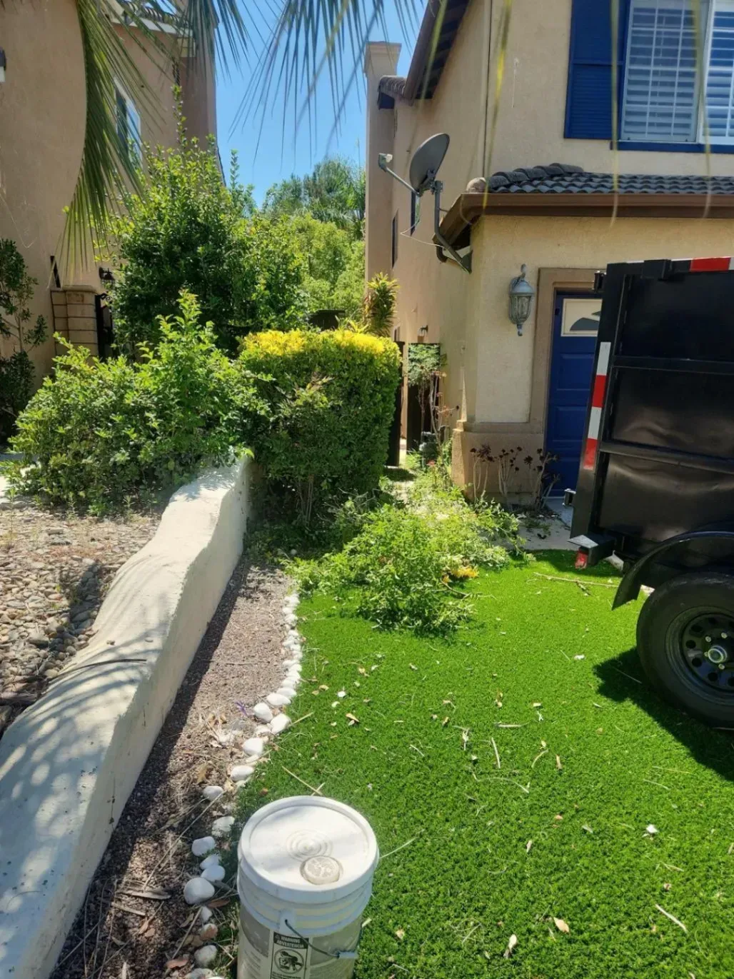 A house with green lawn, trimmed bushes, and a dumpster. Blue door visible. Sunny day.
