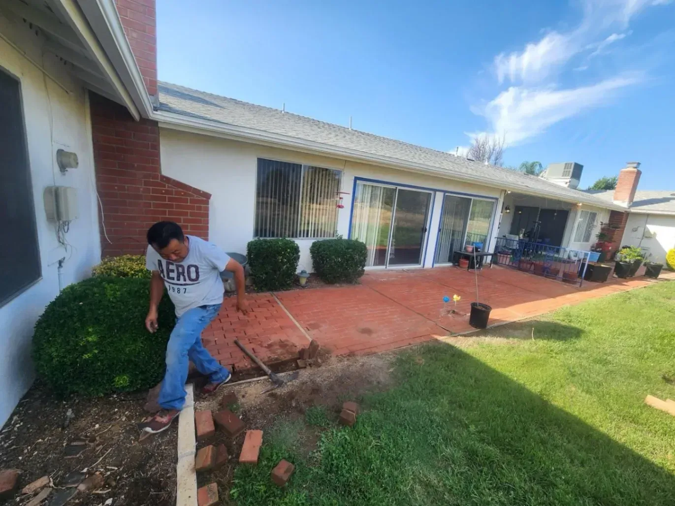 Man laying bricks on a red patio next to a house with a green lawn and blue sky.