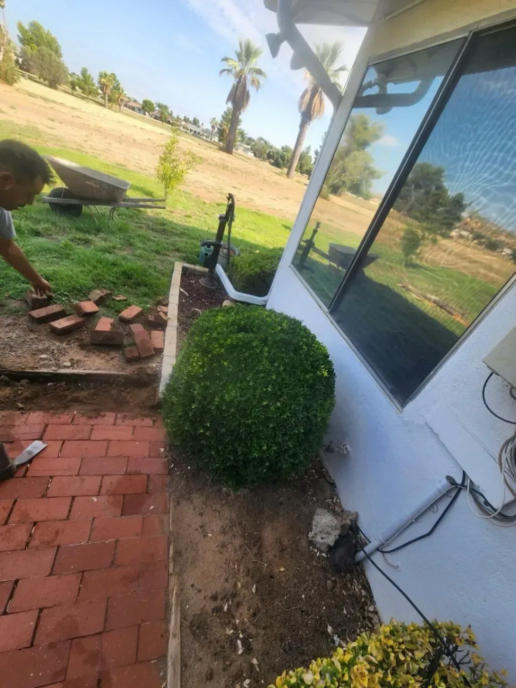 Person laying bricks next to a trimmed green bush. Side of a house with a window and a grassy hillside in the background.