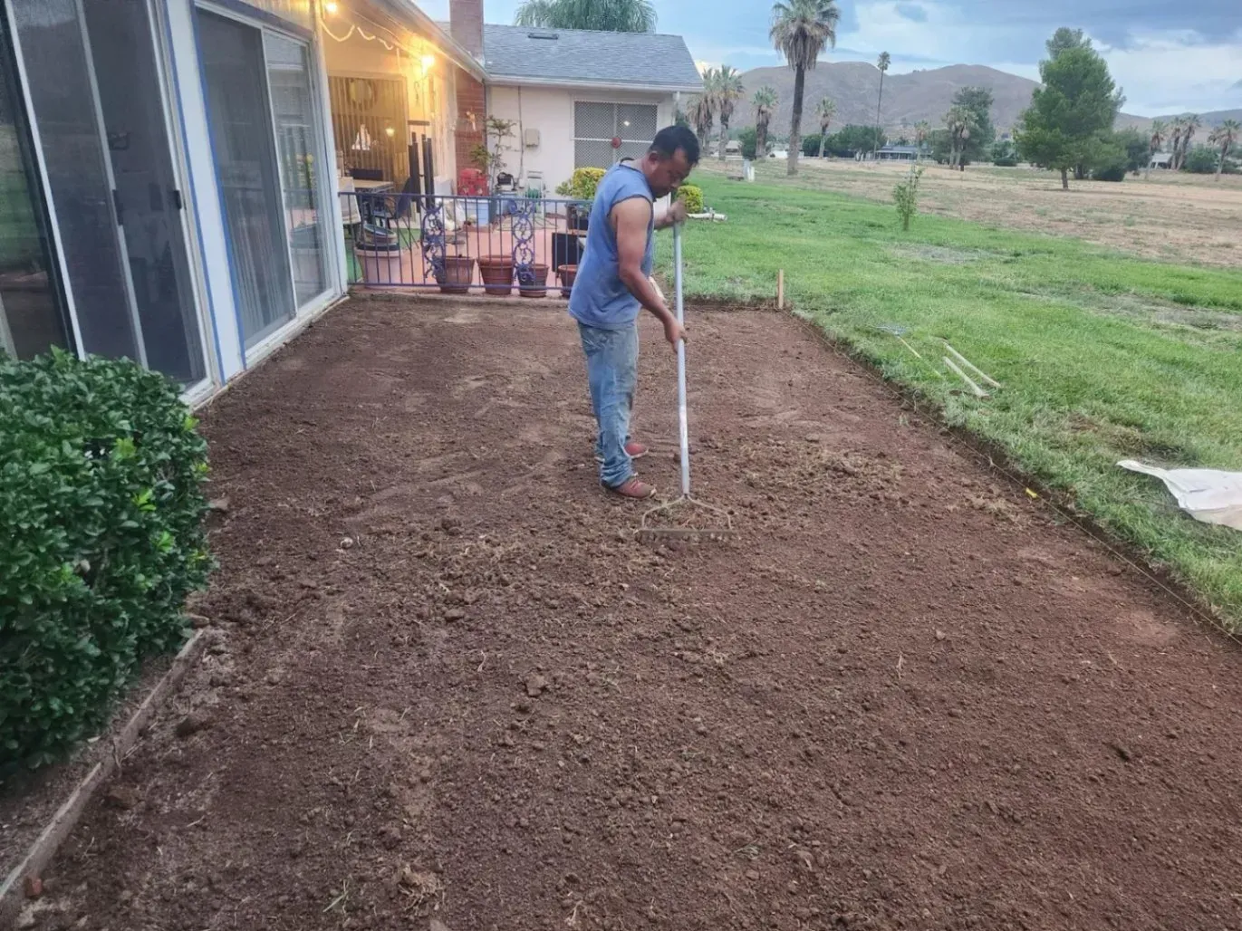 Person raking soil on a patio, preparing the ground for planting. House and yard in the background.