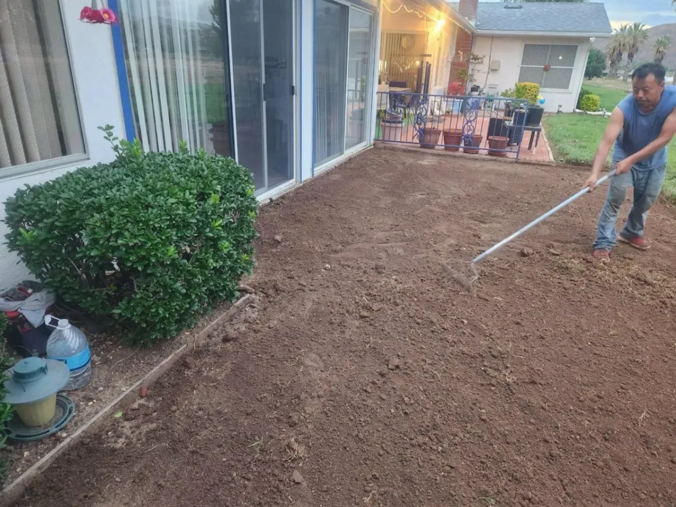 Man rakes soil on patio near a bush and house.
