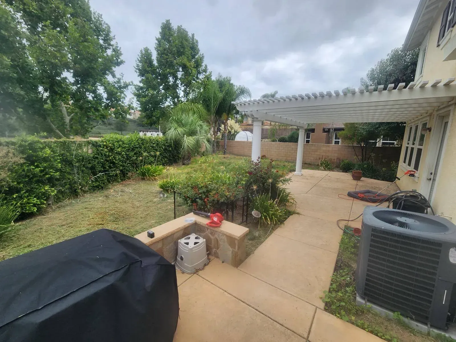 Backyard patio with pergola, grill, air conditioner, and overgrown plants under cloudy sky.