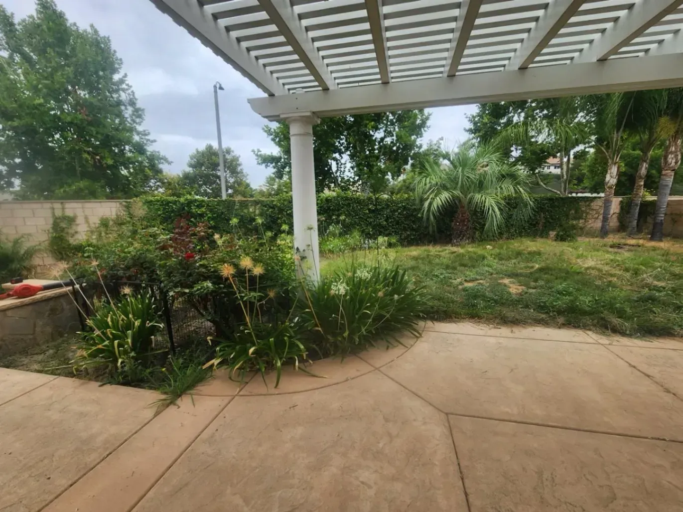 Patio with a pergola, flower bed, and grassy lawn on a cloudy day.