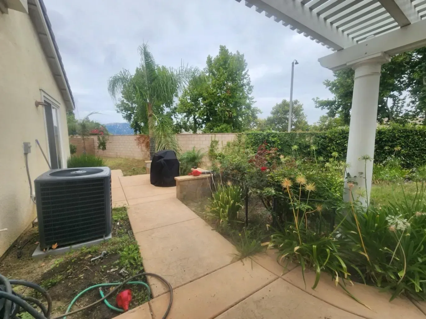 Backyard patio with AC unit, BBQ, flowers, and pergola. Cloudy sky, beige building.