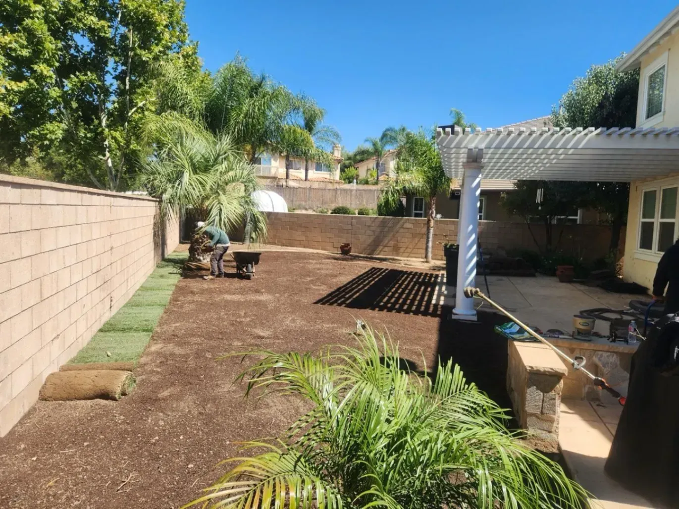 Backyard landscaping with new sod rolls, dirt, and a pergola.