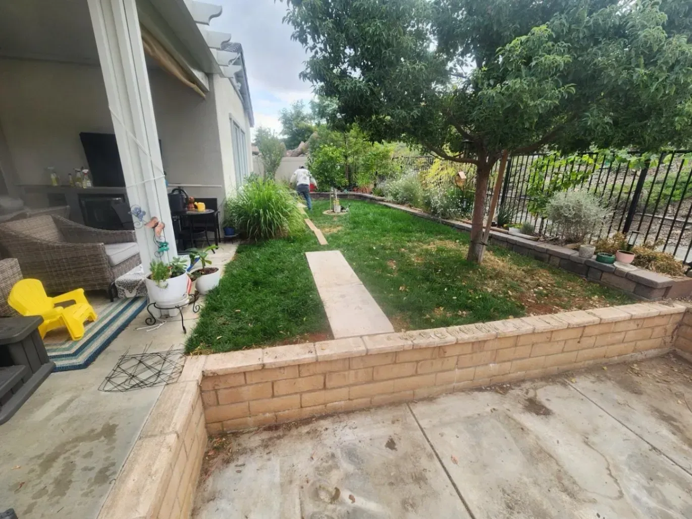 Backyard with patio, lawn, trees, and a brick retaining wall. Person walks on the grass path.