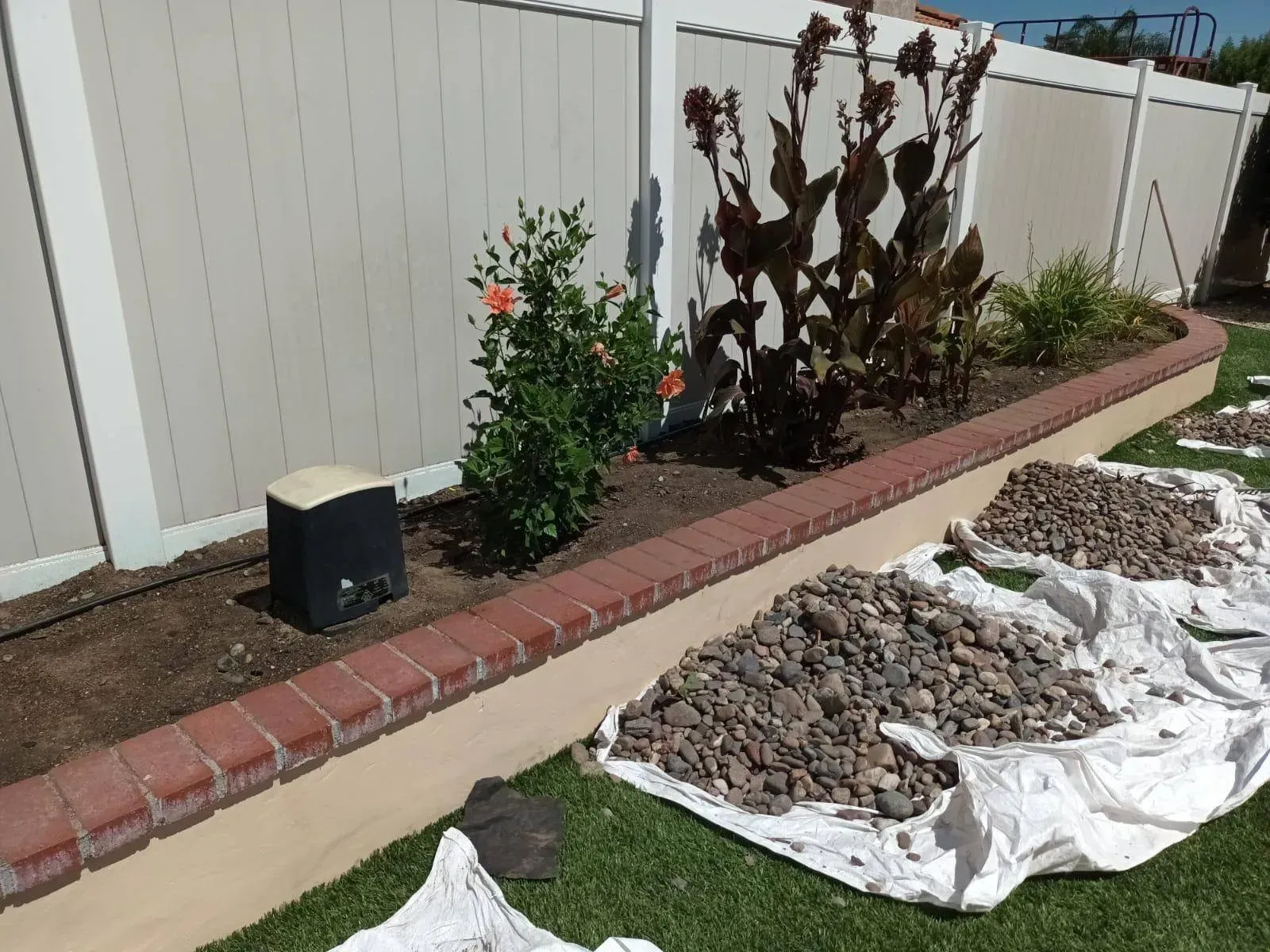 Landscaped flower bed with red brick border, against a white fence. Bags of rocks lay on the grass.