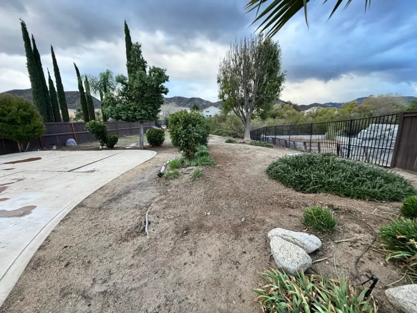 Backyard with dirt and rocks, a concrete path, and various trees and bushes under a cloudy sky.