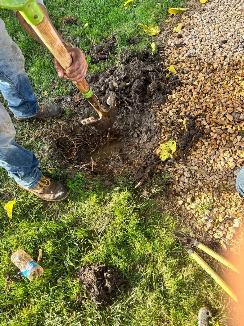 Person digging in the dirt with a shovel, near grass and gravel.