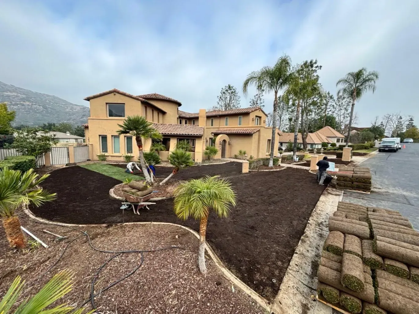 Workers installing sod in front of a tan two-story house with palm trees, a cloudy sky, and a mountain in the background.