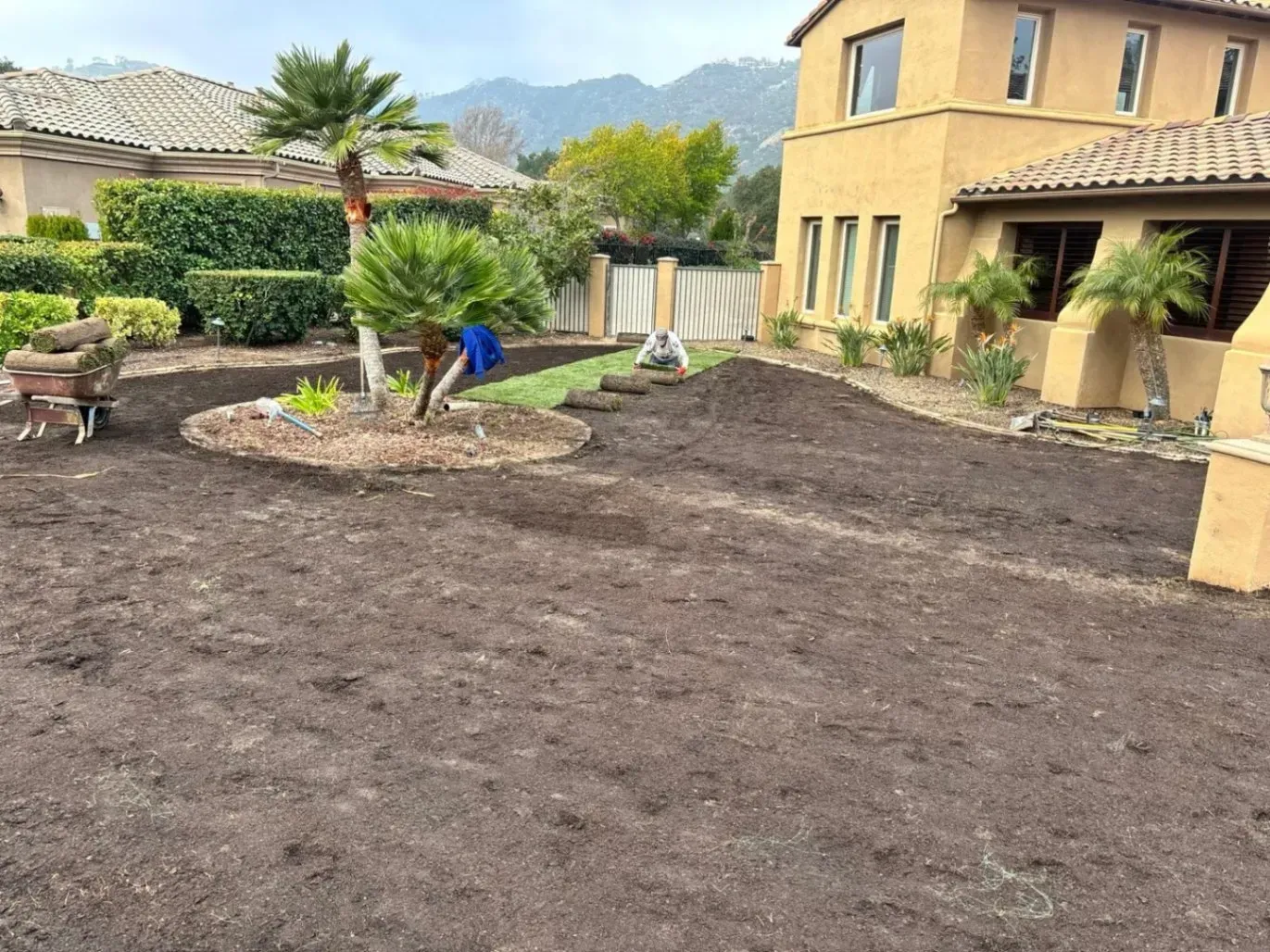 A yard with fresh soil, prepared for landscaping, next to a two-story beige house and mountains in the background.