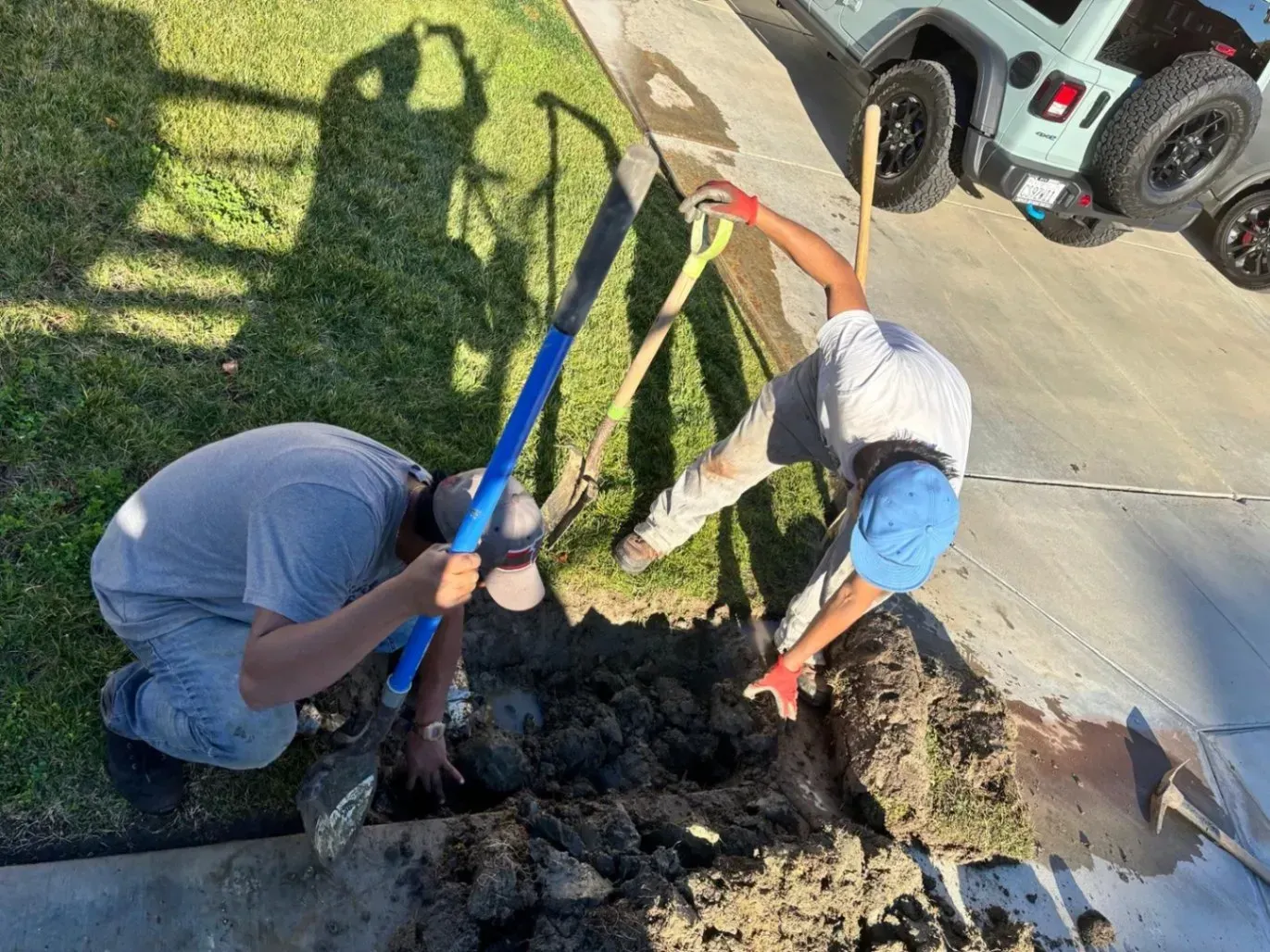 Two people digging in dirt near a sidewalk, one using a shovel and one using a blue-handled tool. A Jeep is parked nearby.
