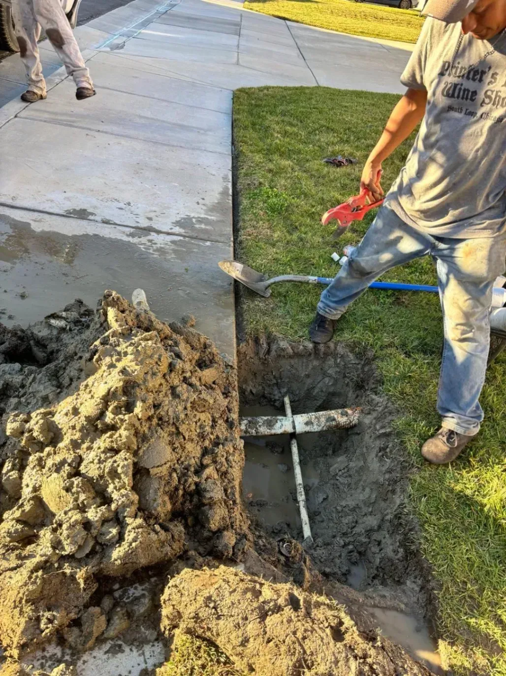 Man digging in a trench by a sidewalk, with exposed pipes and a pile of dirt.