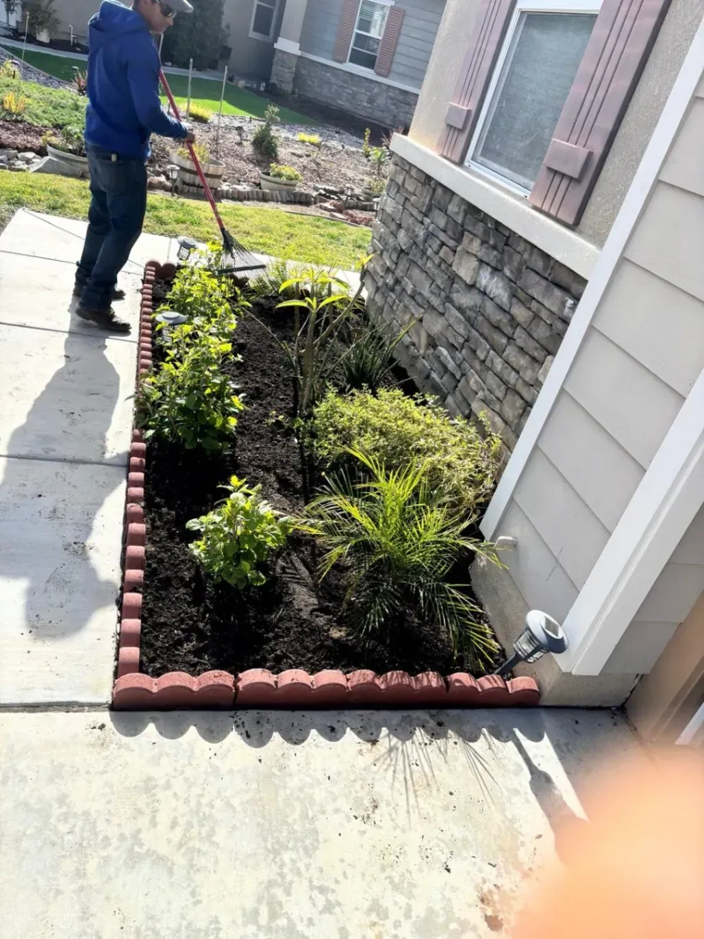Person raking a mulched garden bed next to a house with beige siding and stone accents.