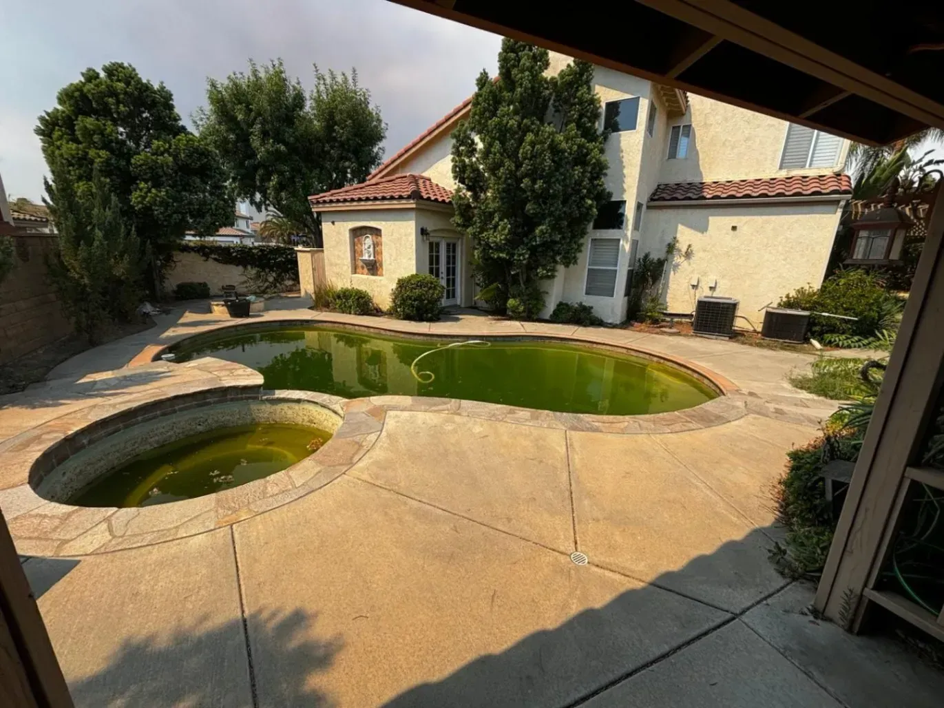 Backyard with a neglected, algae-filled swimming pool and spa, concrete patio, and two-story house.