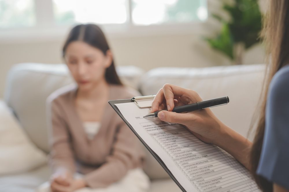 A woman is sitting on a couch while a woman writes on a clipboard.