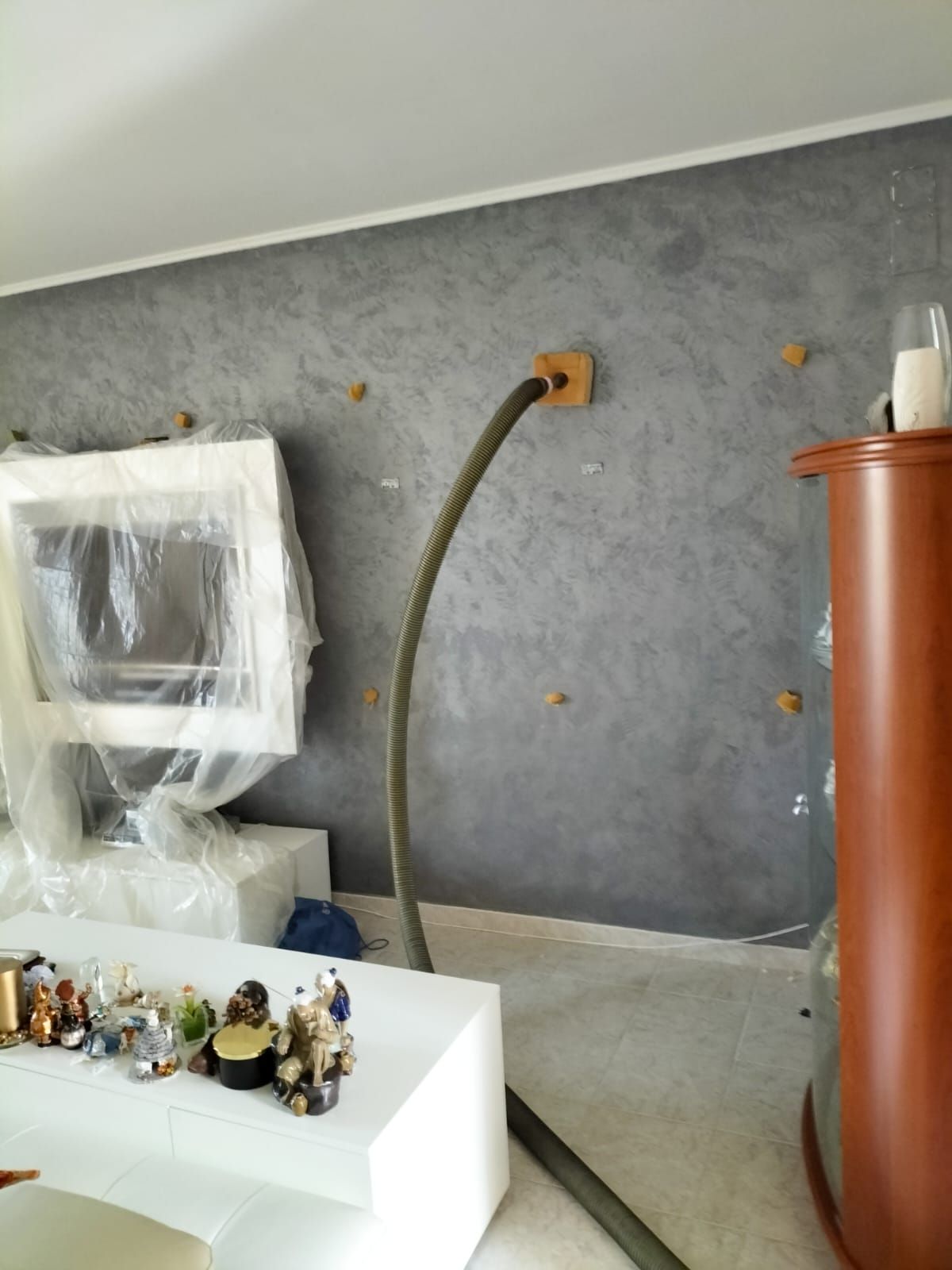 Una parete grigia con una superficie ruvida, dotata di un tubo flessibile centrale collegato a un supporto quadrato in legno e a diversi piccoli tasselli circolari da muro.