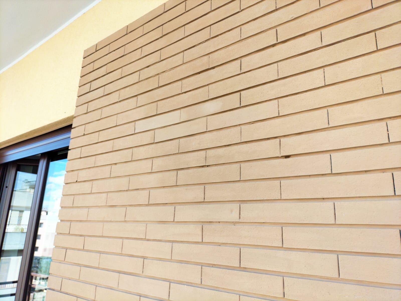 Una sezione di muro in mattoni beige su un balcone accanto a una porta a vetri con telaio in metallo scuro.