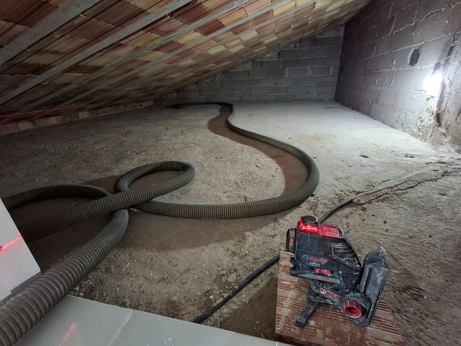 Un tubo flessibile industriale giace sul pavimento polveroso di una soffitta, accanto a un macchinario, sotto un tetto spiovente.