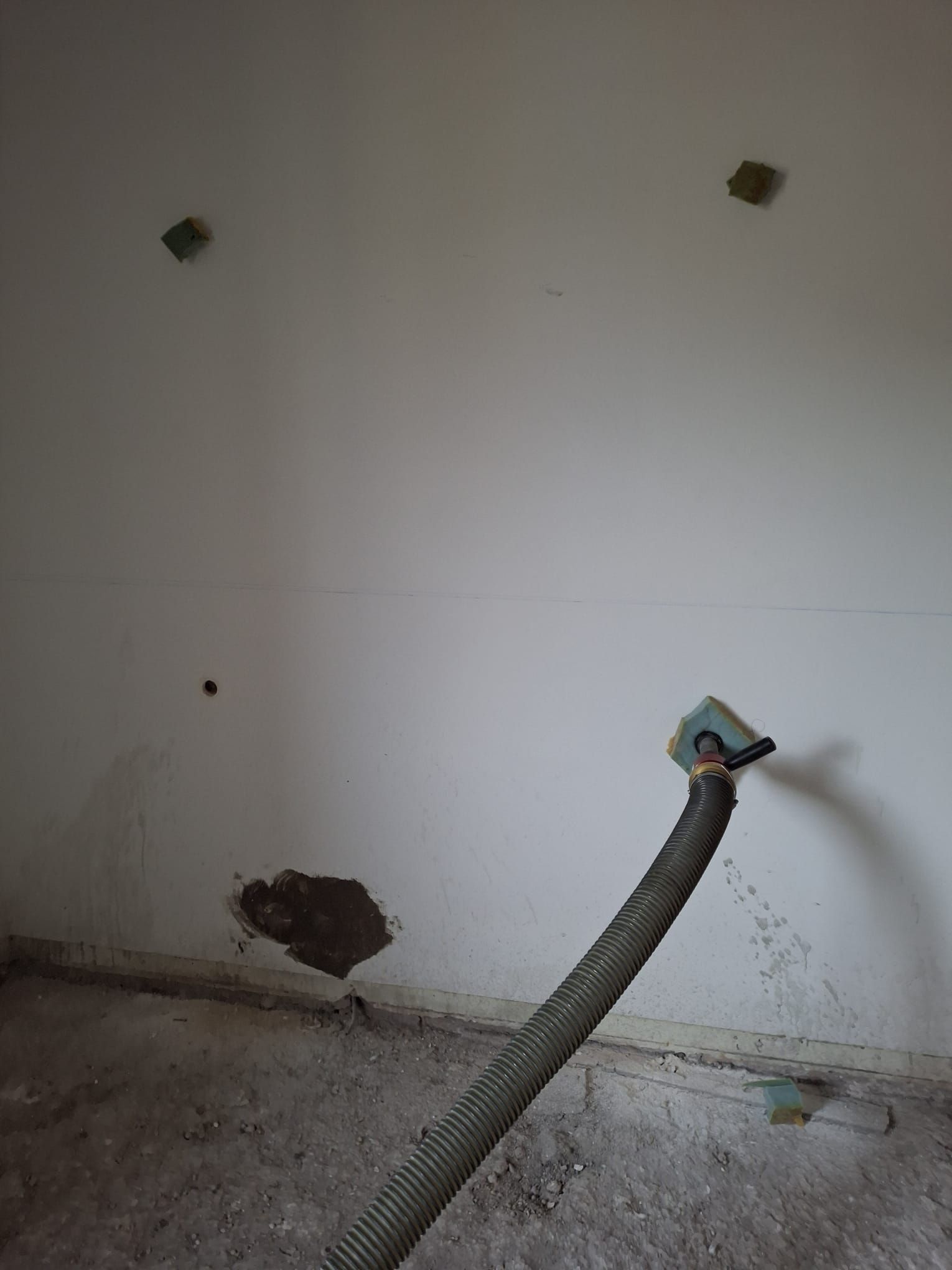 Una stanza non rifinita con pareti bianche, due piccole plafoniere, una porzione di cemento a vista e un tubo di scarico flessibile.