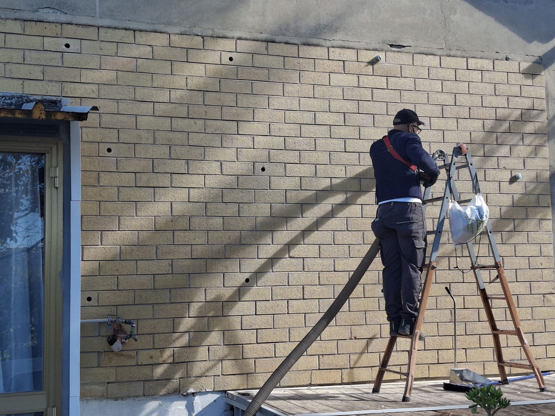 Una persona su una scala lavora su un muro esterno di mattoni chiari, praticando dei fori nella muratura.