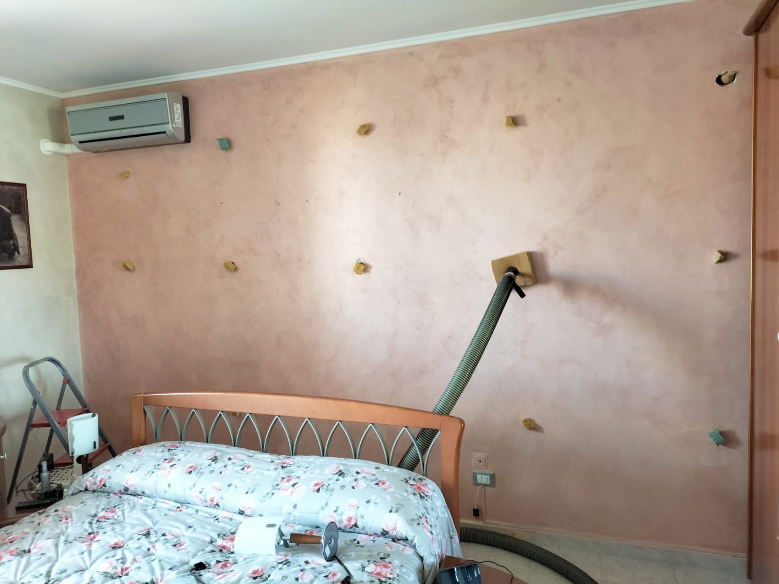 Parete della camera da letto con pittura rosa a effetto materico e piccoli ganci a muro, con un tubo flessibile che sporge da una tavola di legno centrale.