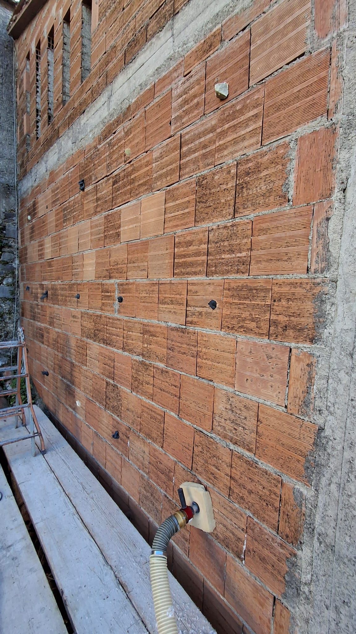 Vista dal basso di un muro di mattoni rossi in costruzione, con un tubo bianco che sporge dalla parte inferiore.