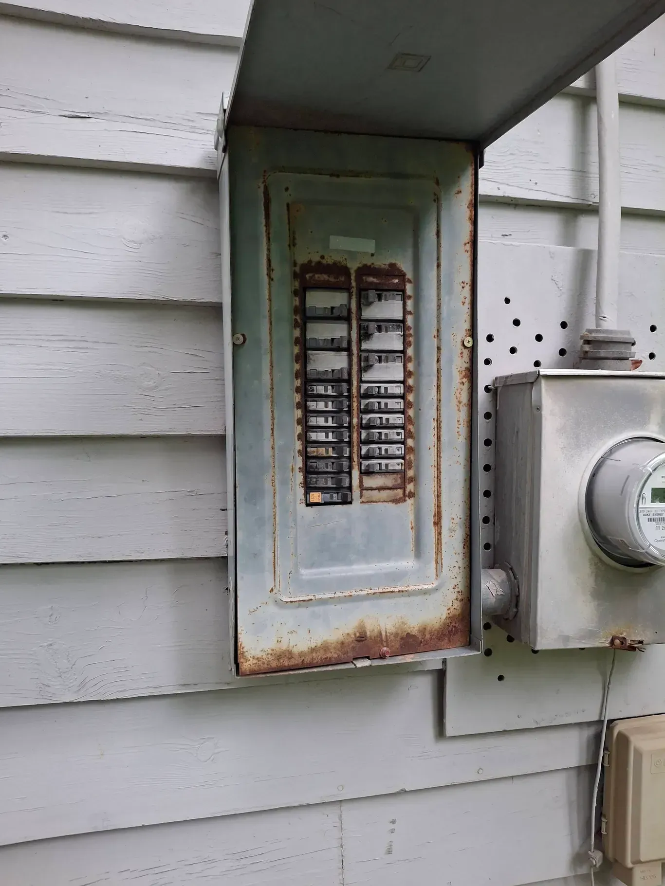 Exterior electrical panel with rust, attached to a house with white siding, next to a meter.