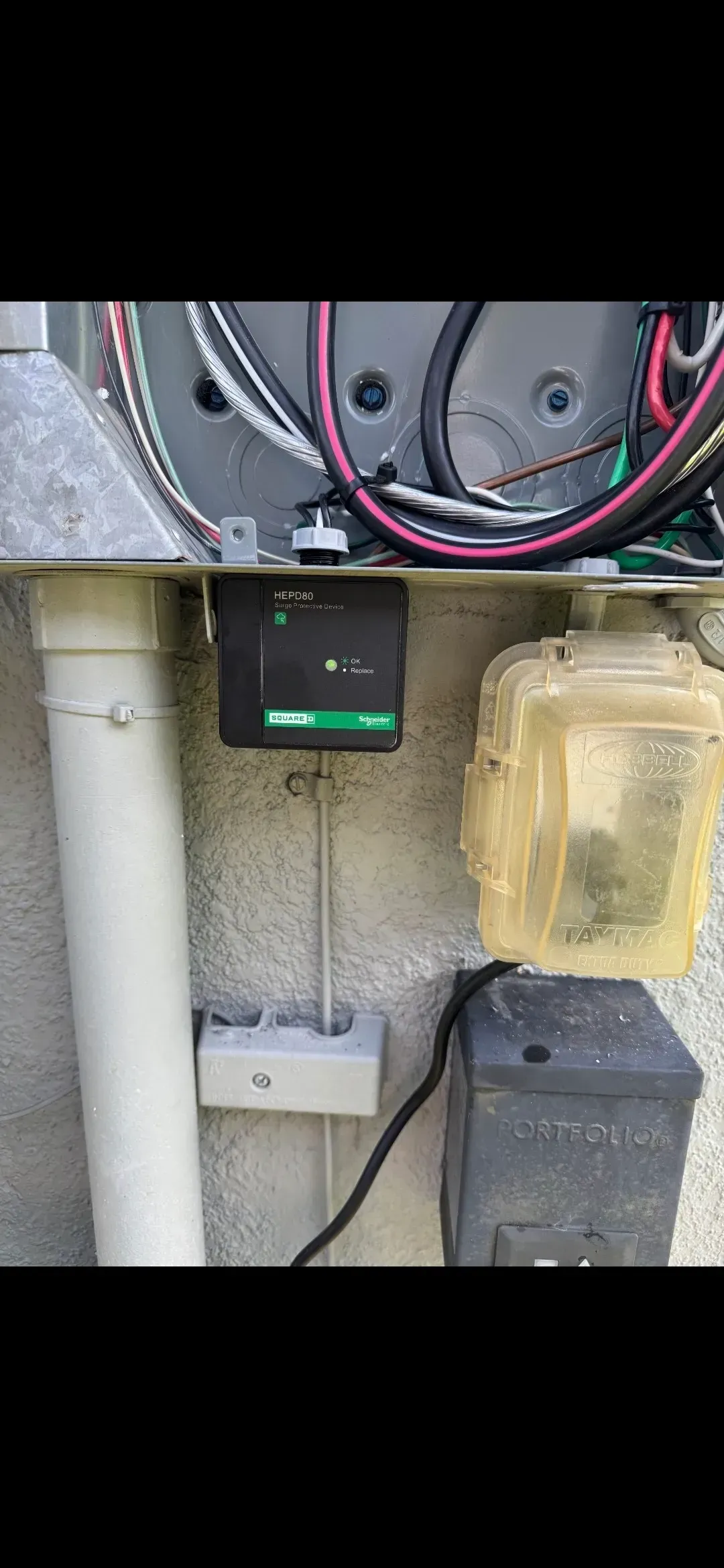 A black box with a green light on a wall, next to a white pipe, outlet, and weatherproof cover.