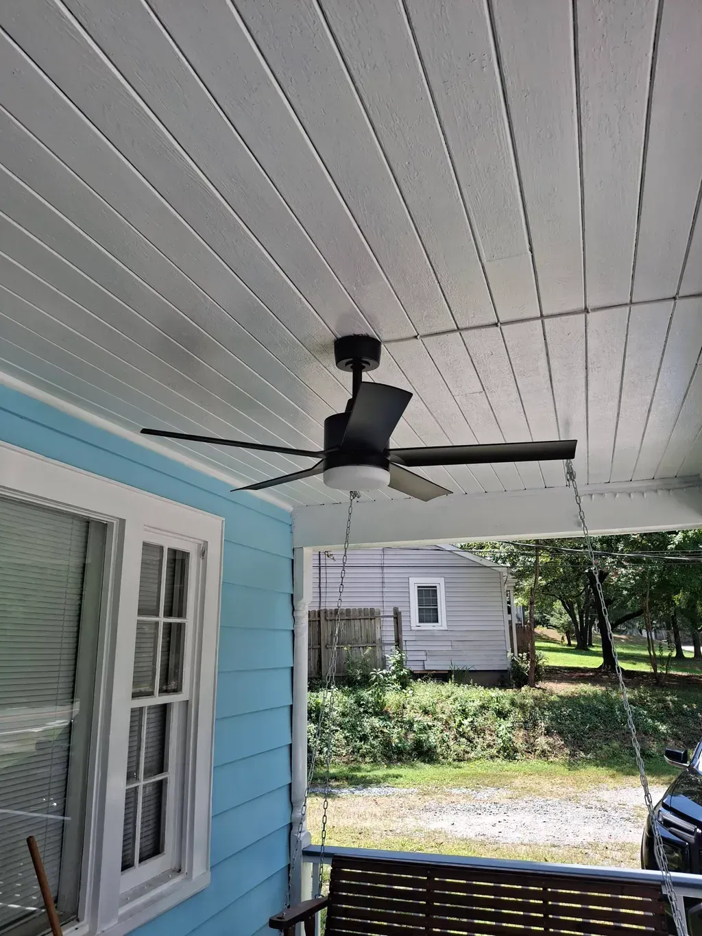 Black ceiling fan with light on a blue porch, with a view of a yard and house.