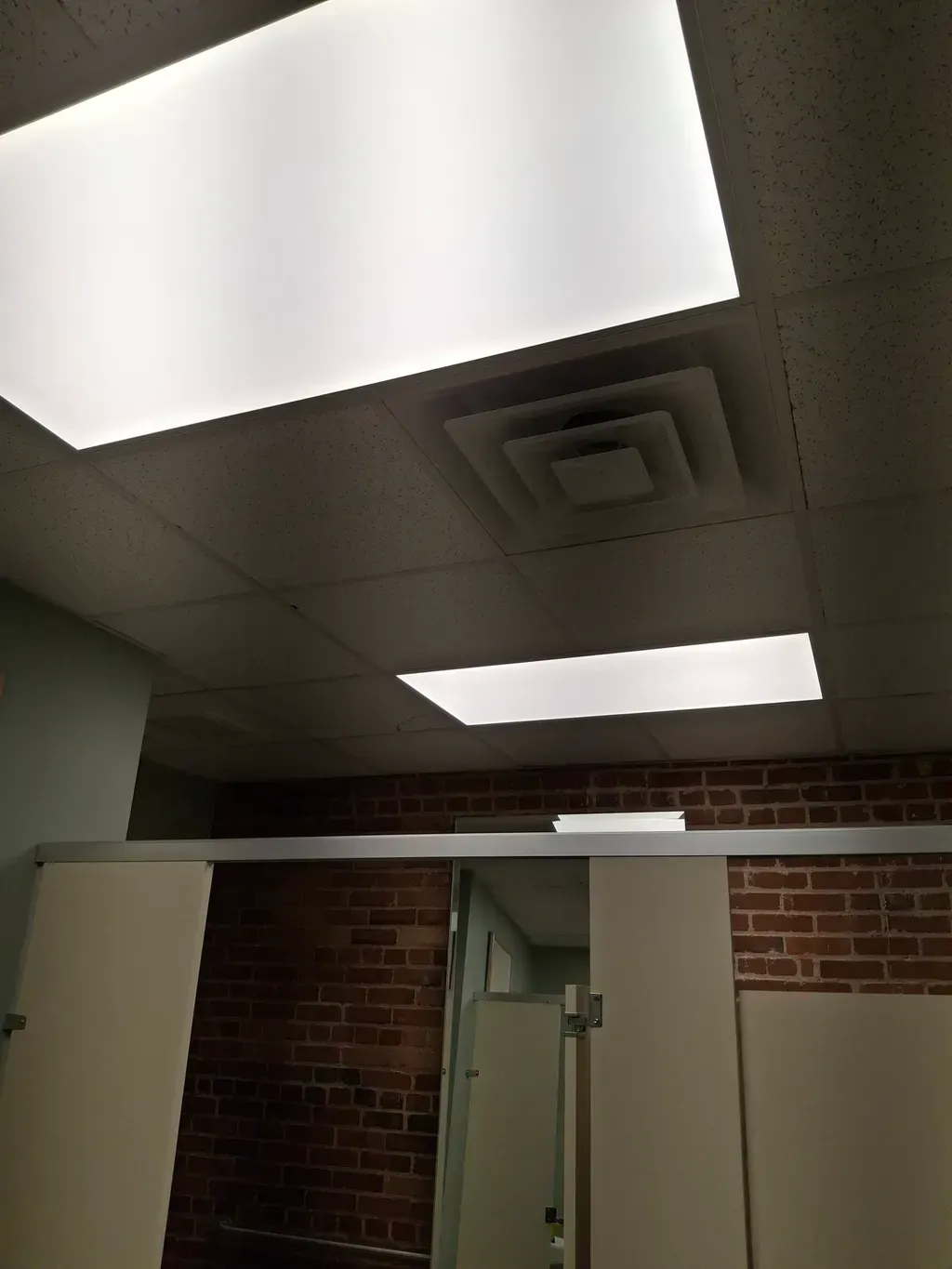 Public restroom ceiling with rectangular lights, air vent, and brick wall. White stall doors are in the foreground.