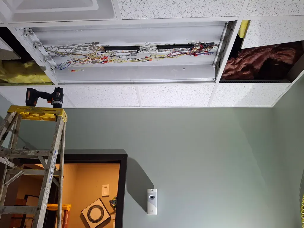 Person on ladder working on ceiling tile and light fixture. Wires exposed; drywall and door in view.