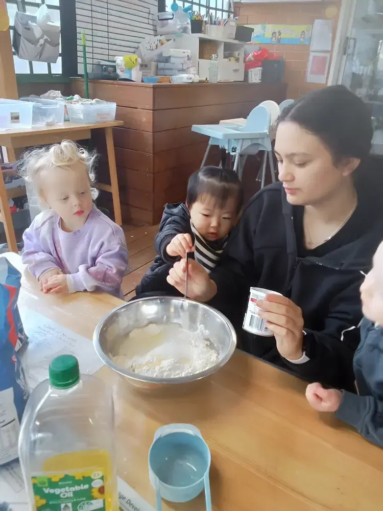 A childcare educator supports a toddler in adding an ingredient to a large mixing bowl during a fun and educational cooking activity.