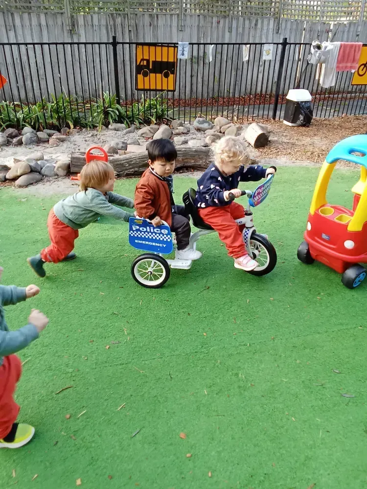 Children at a childcare enjoy outdoor playtime on artificial grass, with one child riding a tricycle, another sitting in the back, and a third child playfully pushing them.