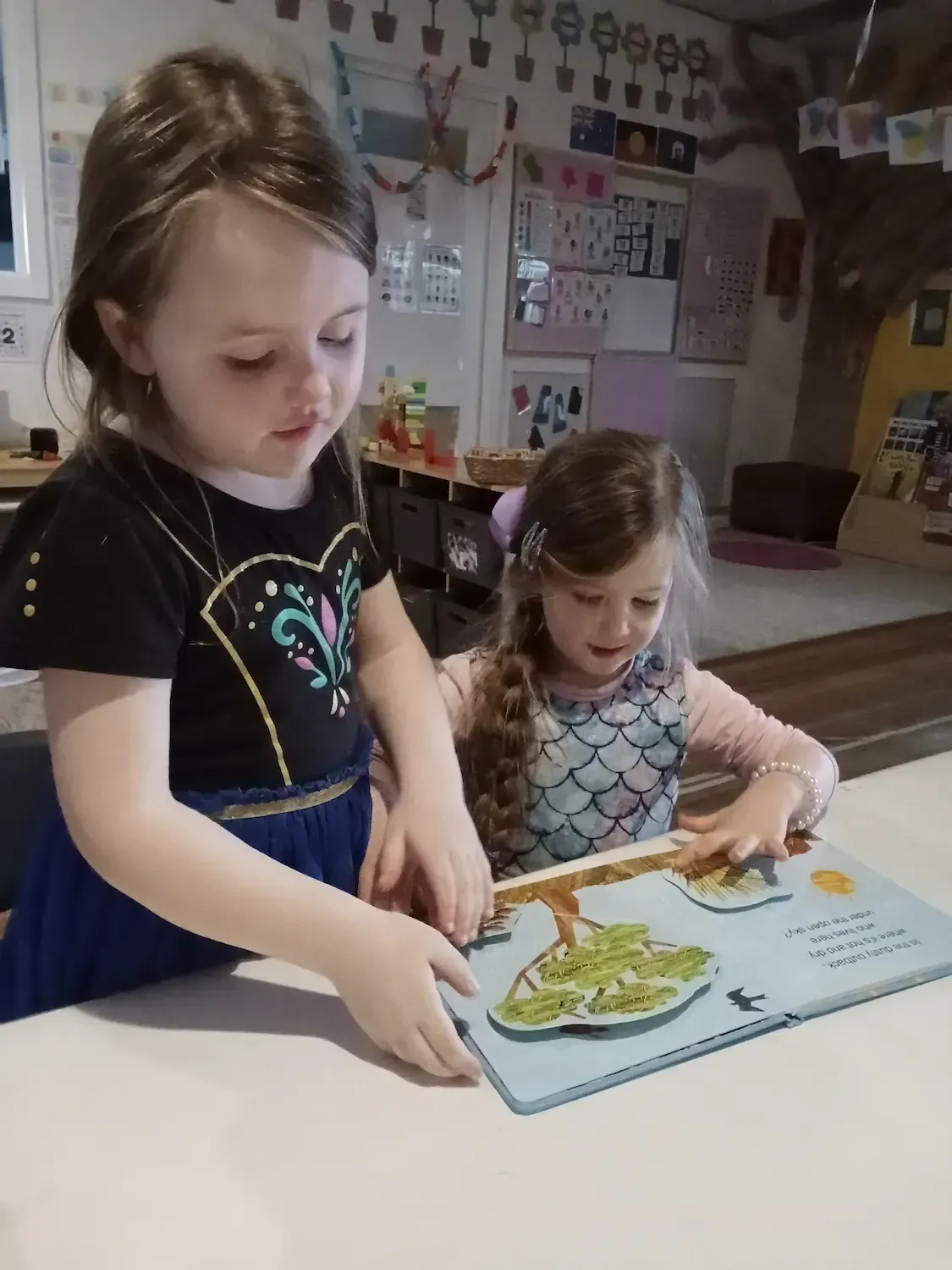 Two young girls at a childcare centre engage in a shared reading activity, exploring a colourful pop-up book together.